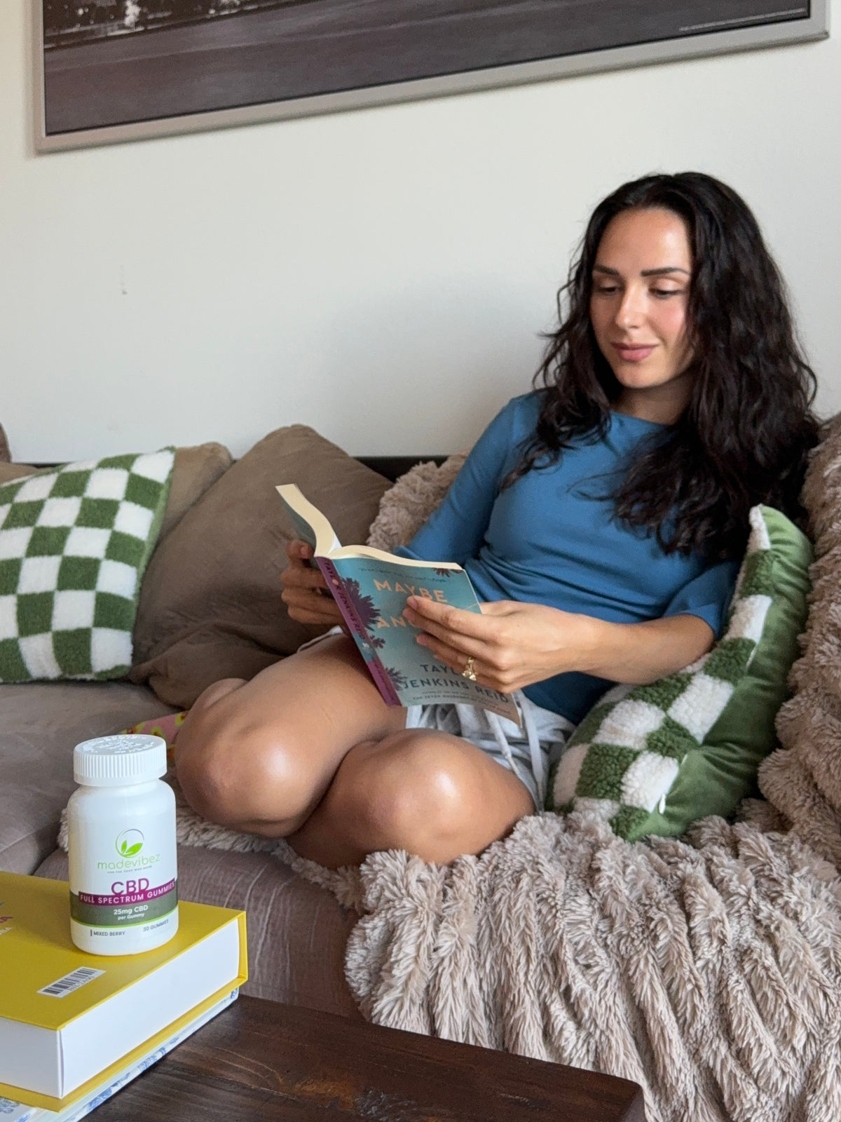 A woman with long dark hair sits on a couch reading a book, wearing a blue long-sleeve top and white shorts. A bottle of CBD oil on a yellow book is visible in the foreground on a table.