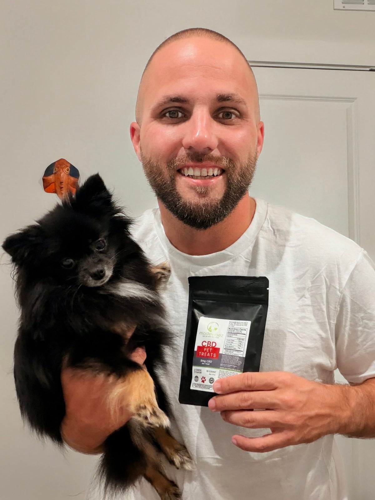 A smiling man in a white t-shirt holds a small black and tan dog over his shoulder while holding up a black stand-up pouch labeled 'CBD Pet Treats' in a home setting.