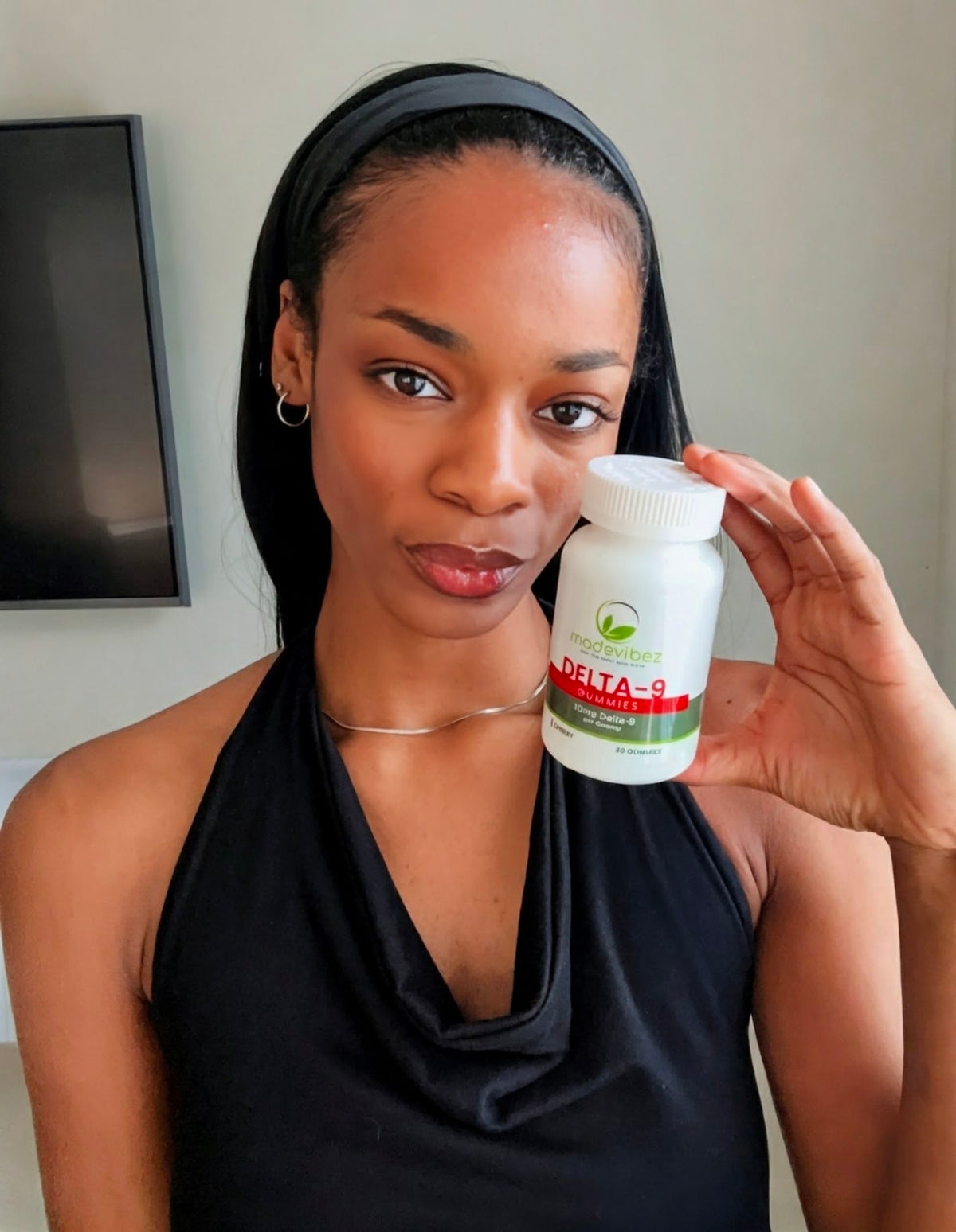 A woman wearing a black halter top and black headband holds up a white bottle of Organic Heroes Delta-9 edibles in front of her face.