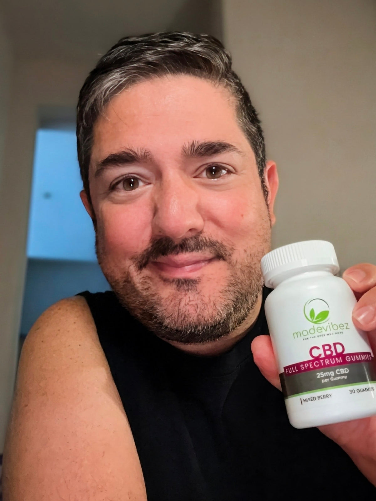 A man holding a white bottle of ModeVibeZ CBD Full Spectrum Gummies next to his face in a selfie-style close-up.