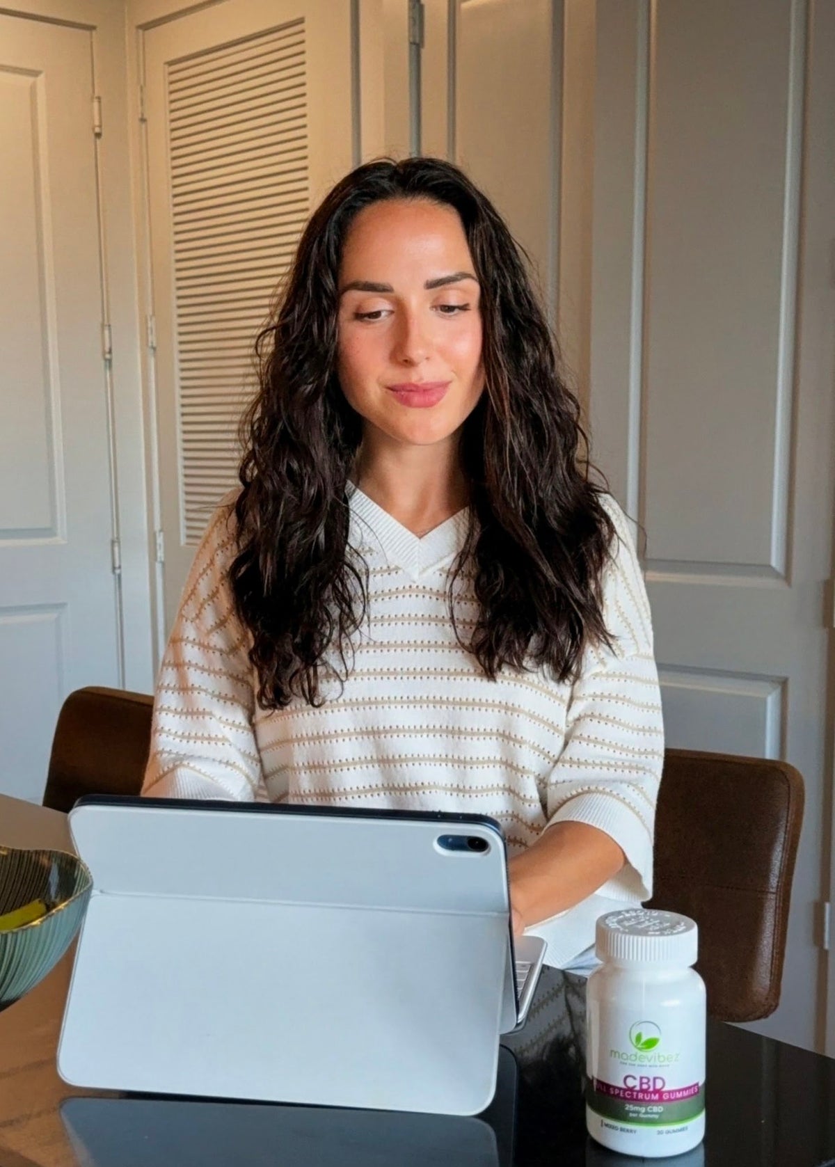 A woman with long dark wavy hair sits at a table using a white tablet, with a white bottle of CBD oil in front of her. She is wearing a cream-colored knit top with thin brown horizontal stripes.
