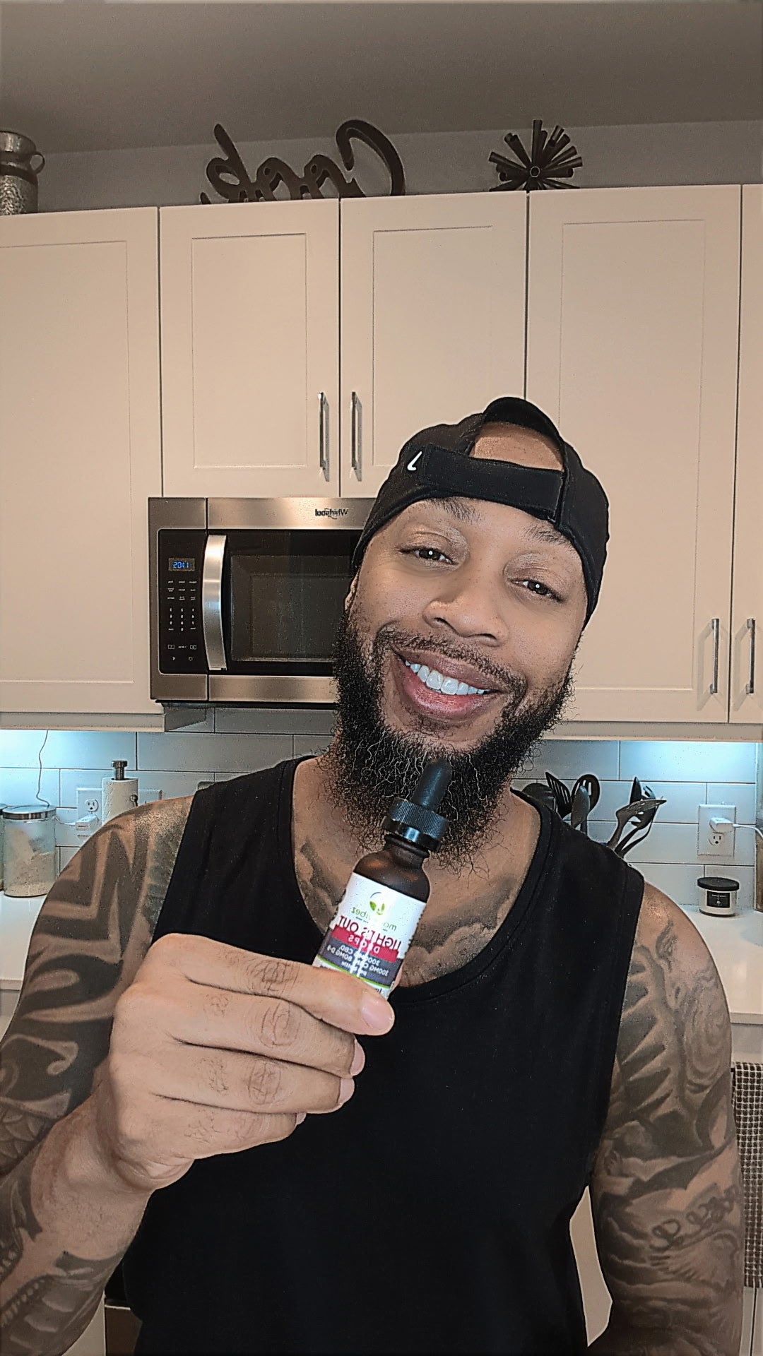 A smiling man with a beard and tattoos wearing a black tank top and backward black cap, holding up a small amber glass dropper bottle in a kitchen setting with white cabinets and a stainless steel microwave behind him.