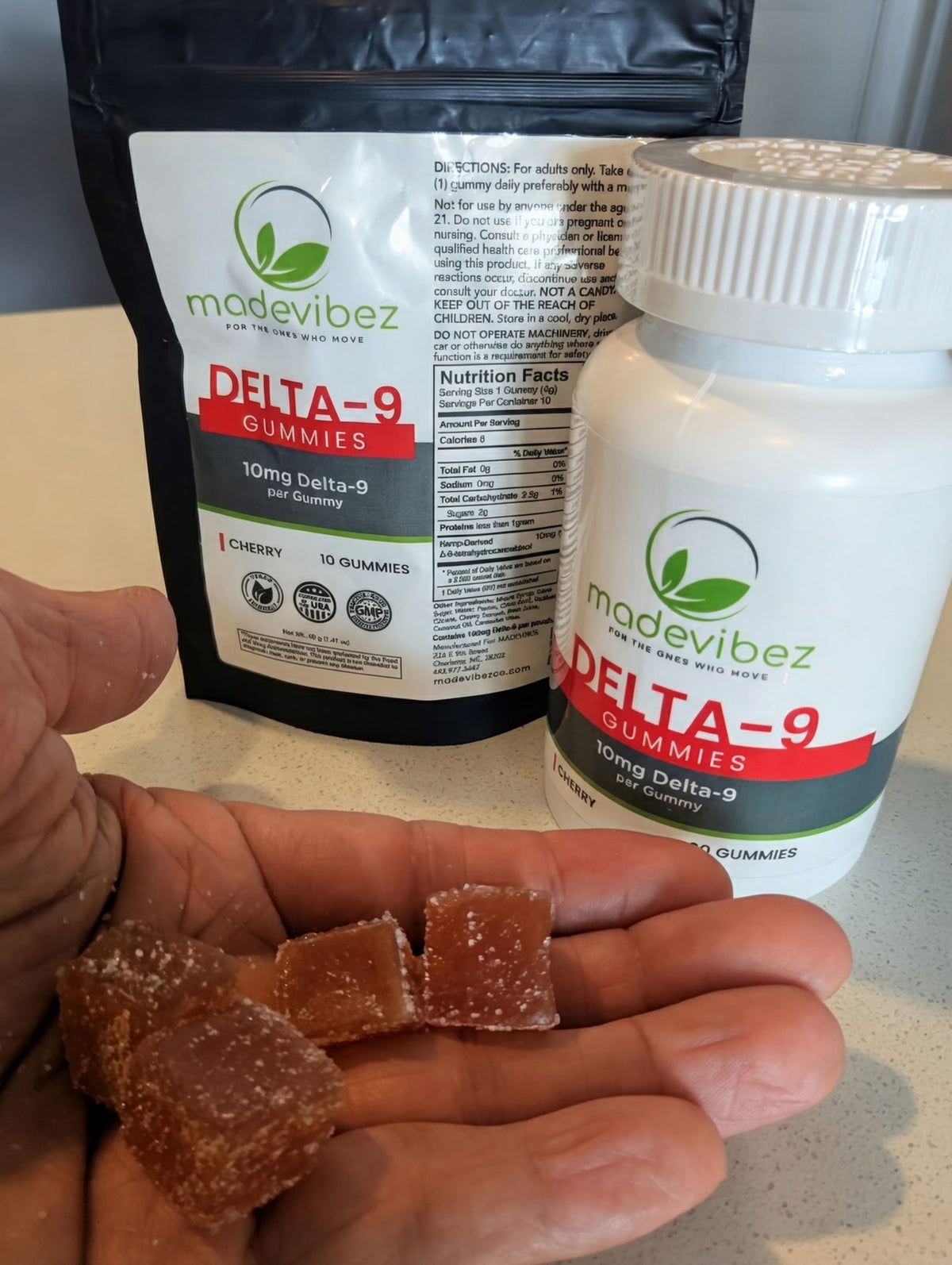 A hand holds several translucent brown gummy candies in front of a white bottle and a black pouch of Madevibez Delta-9 Gummies on a kitchen counter.