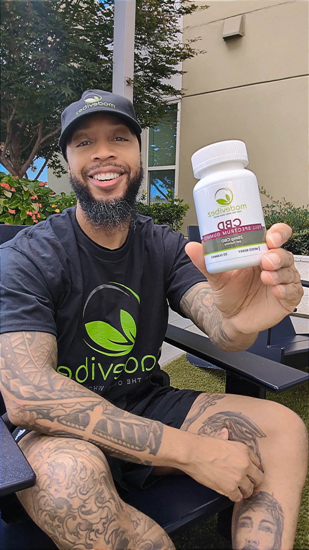 A smiling man wearing a black t-shirt and cap sits outdoors holding up a white bottle of 'MediGrown' CBD + Omega 3 supplement. The bottle label features green and purple branding.