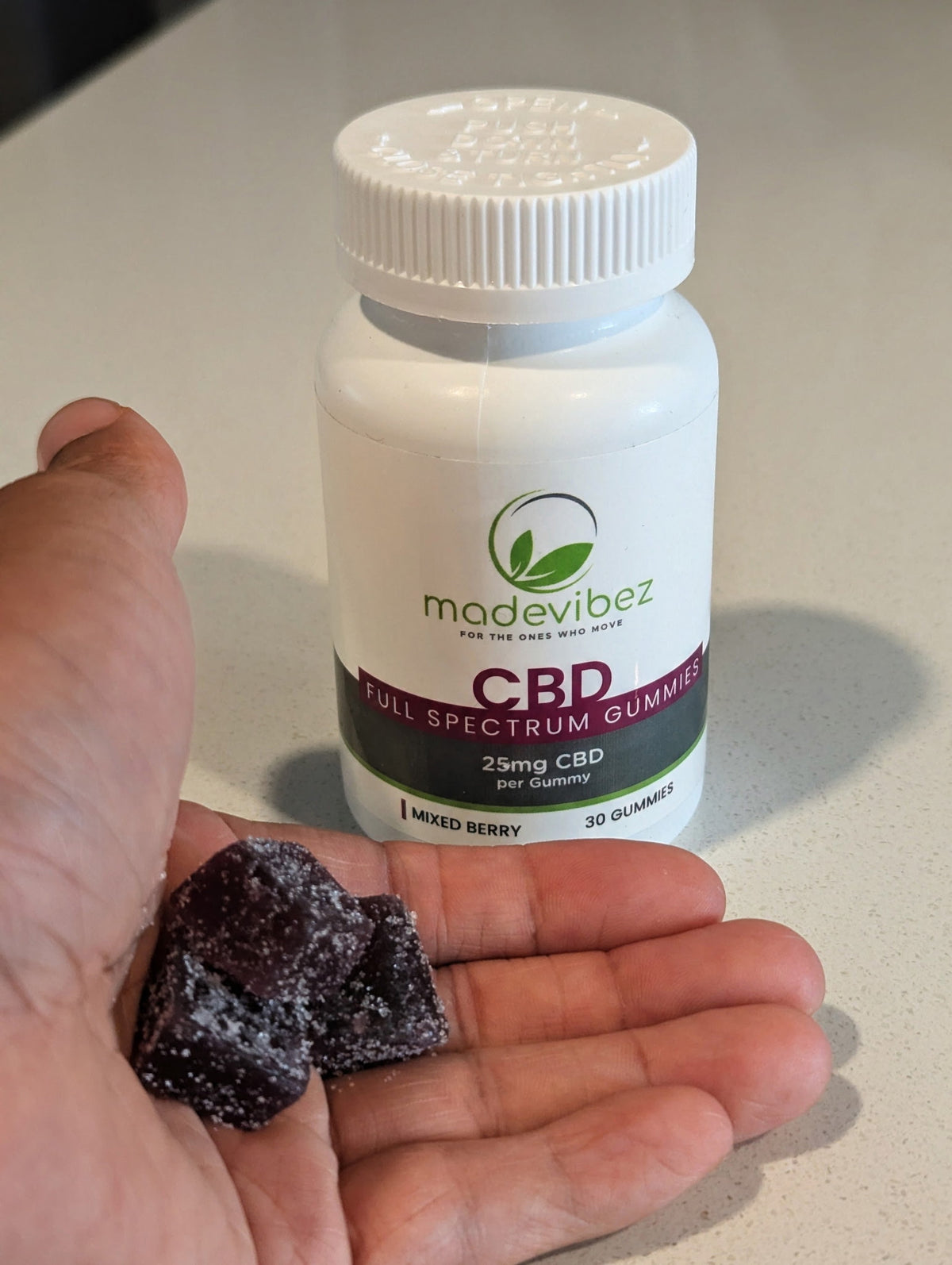 A hand holding two dark gummy candies in front of a white bottle of madevibeZ CBD Full Spectrum Gummies on a light countertop.