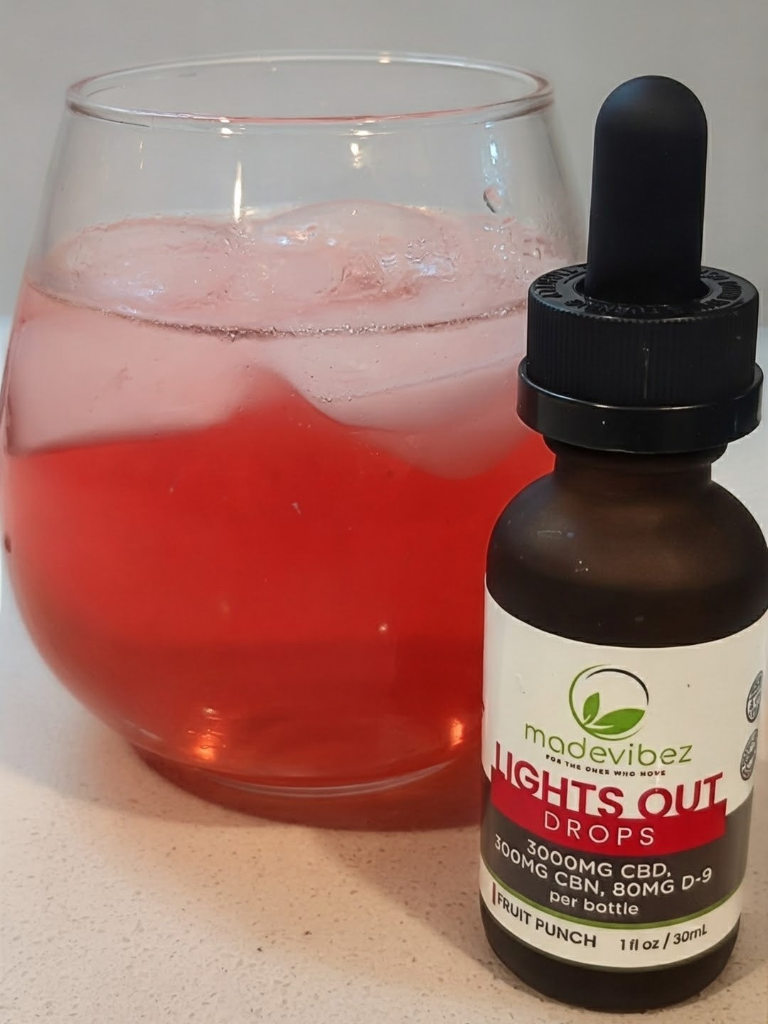 A bottle of MadeVibez Lights Out Drops CBD tincture with a black dropper cap next to a glass of pinkish-red drink with ice on a light countertop.