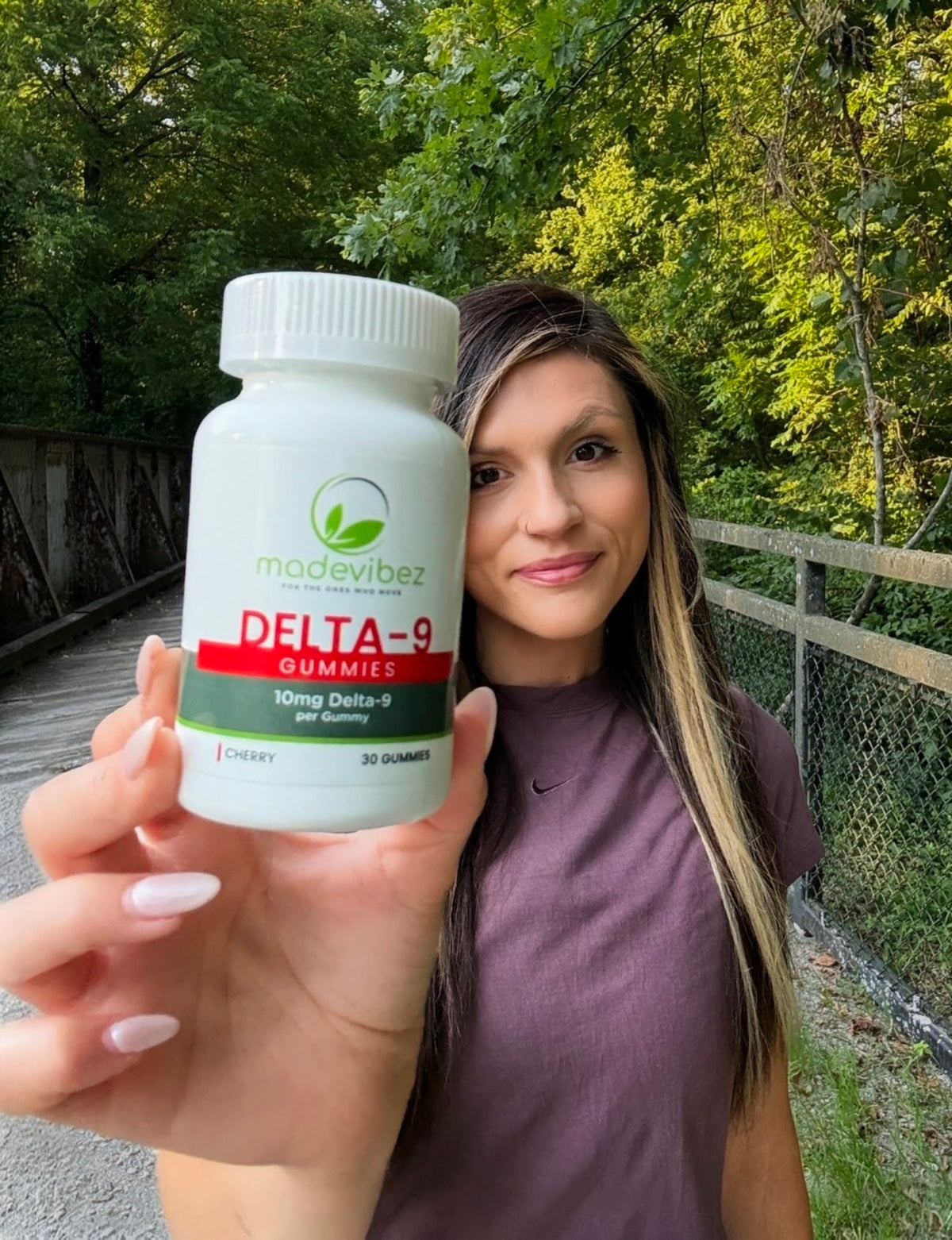 A woman outdoors holds up a white bottle of Madevitez Delta-9 Gummies, with a green forest and fence in the background.
